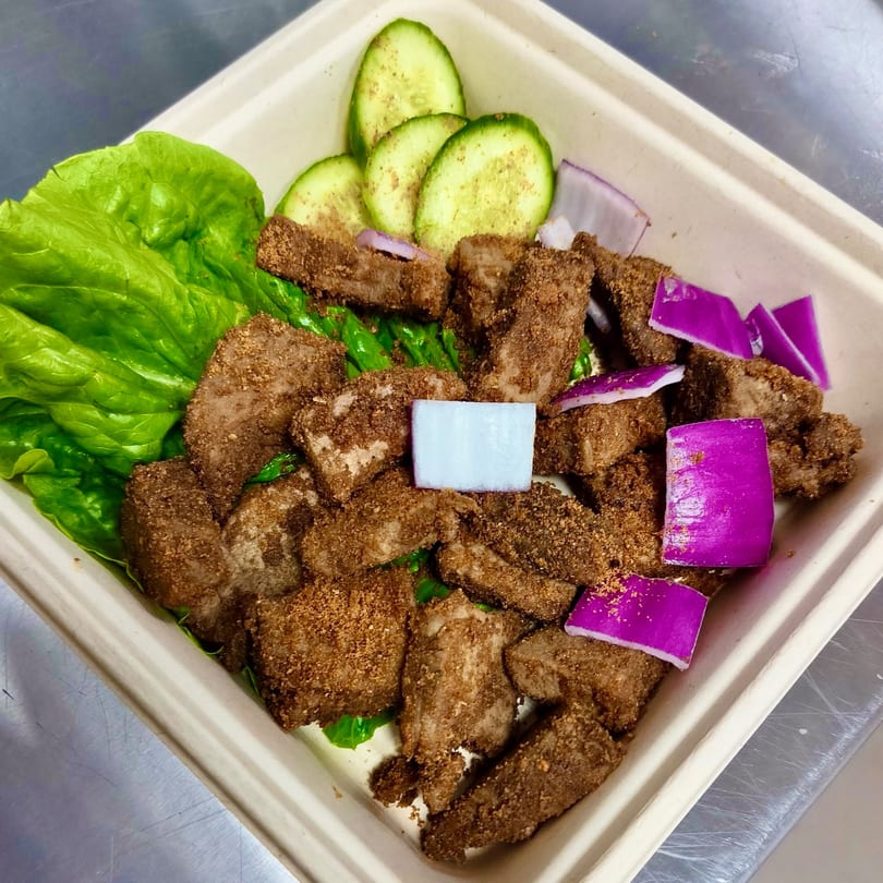Suya Outpost's African-fusion dish with seasoned meat, lettuce, cucumber slices, and red onion in a takeout container.