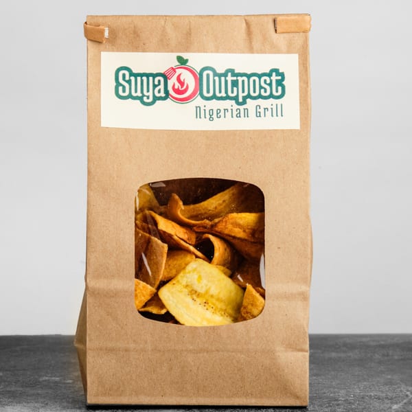 A brown paper bag of Suya Outpost plantain chips with a clear window showing the chips.