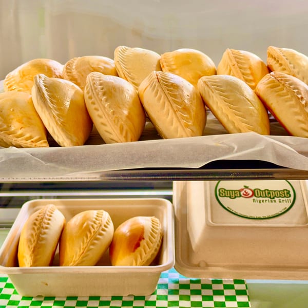 Rows of golden-brown Nigerian meat pies on a tray, with a takeout container from Suya Outpost below.
